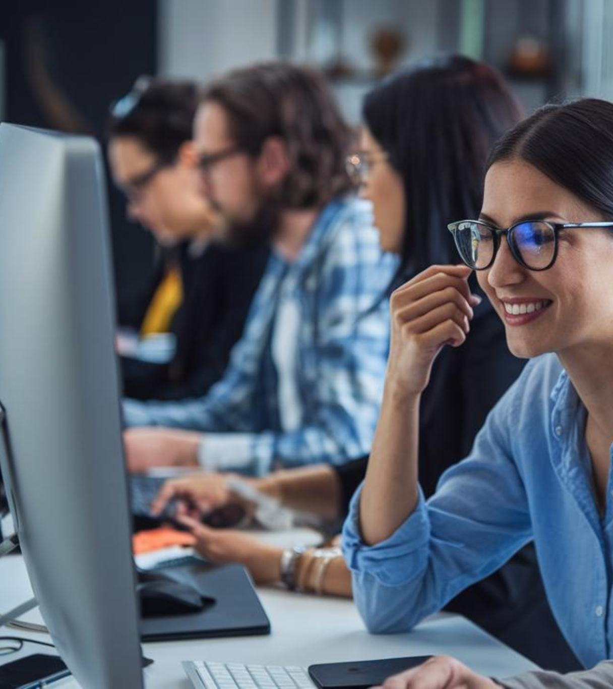 Primer plano de una mujer con gafas y camisa azul sonriendo mientras trabaja en una computadora. Detrás de ella, otros compañeros de trabajo están concentrados en sus tareas en un ambiente colaborativo.